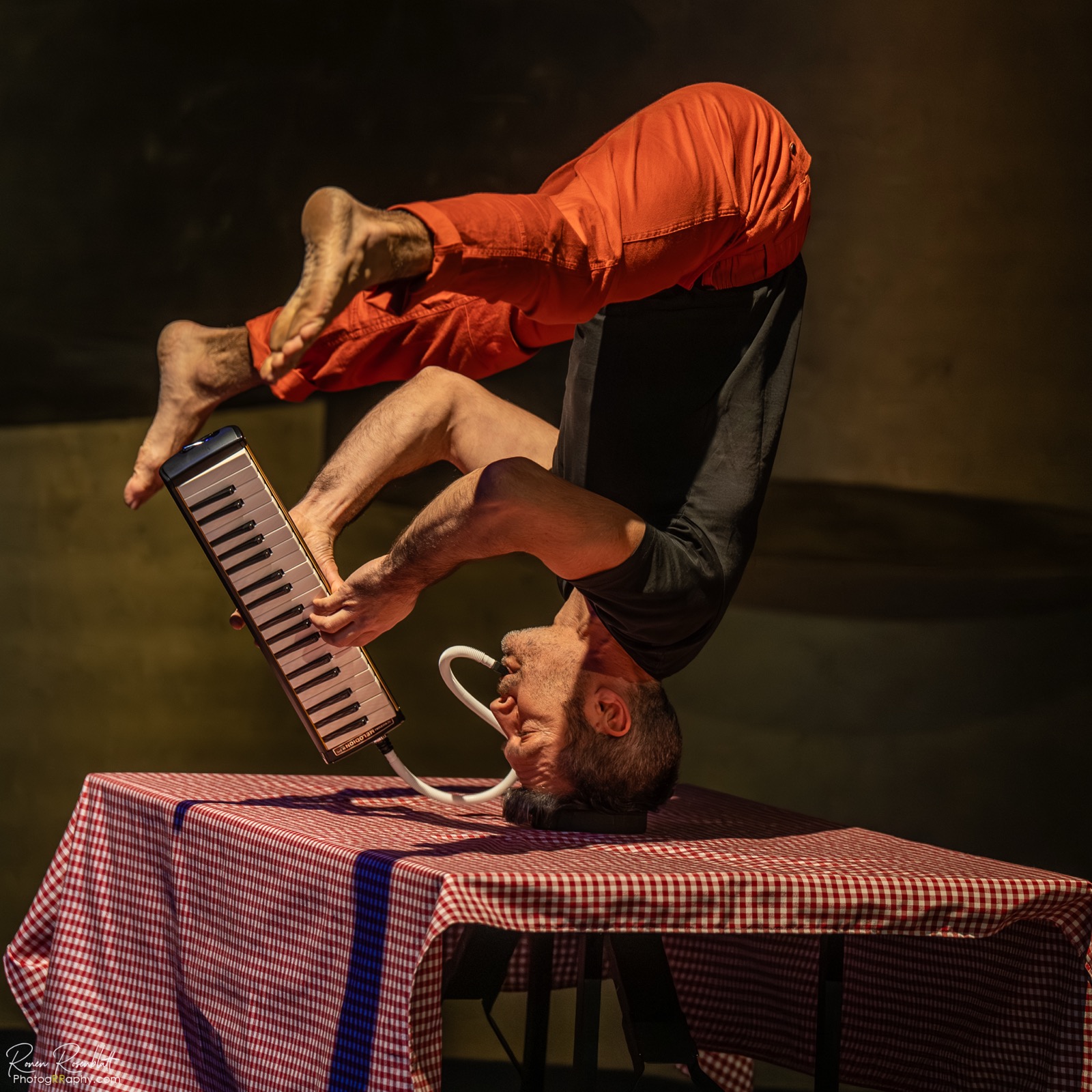 Yuval Oz balancing on his head on a table while playing the melodica during a performance