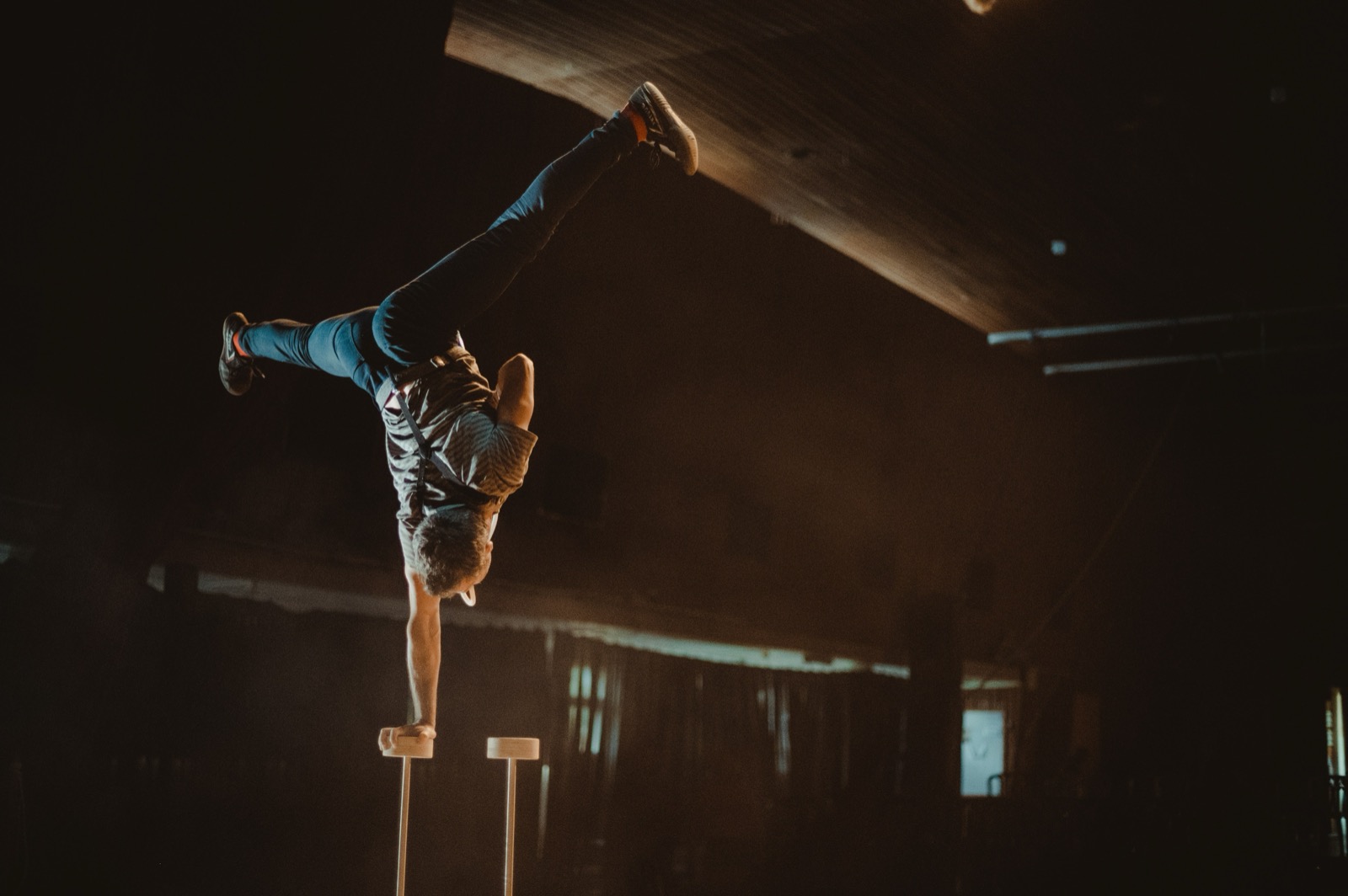 Yuval Oz performing a one-arm handstand on wooden canes in a dark performance space